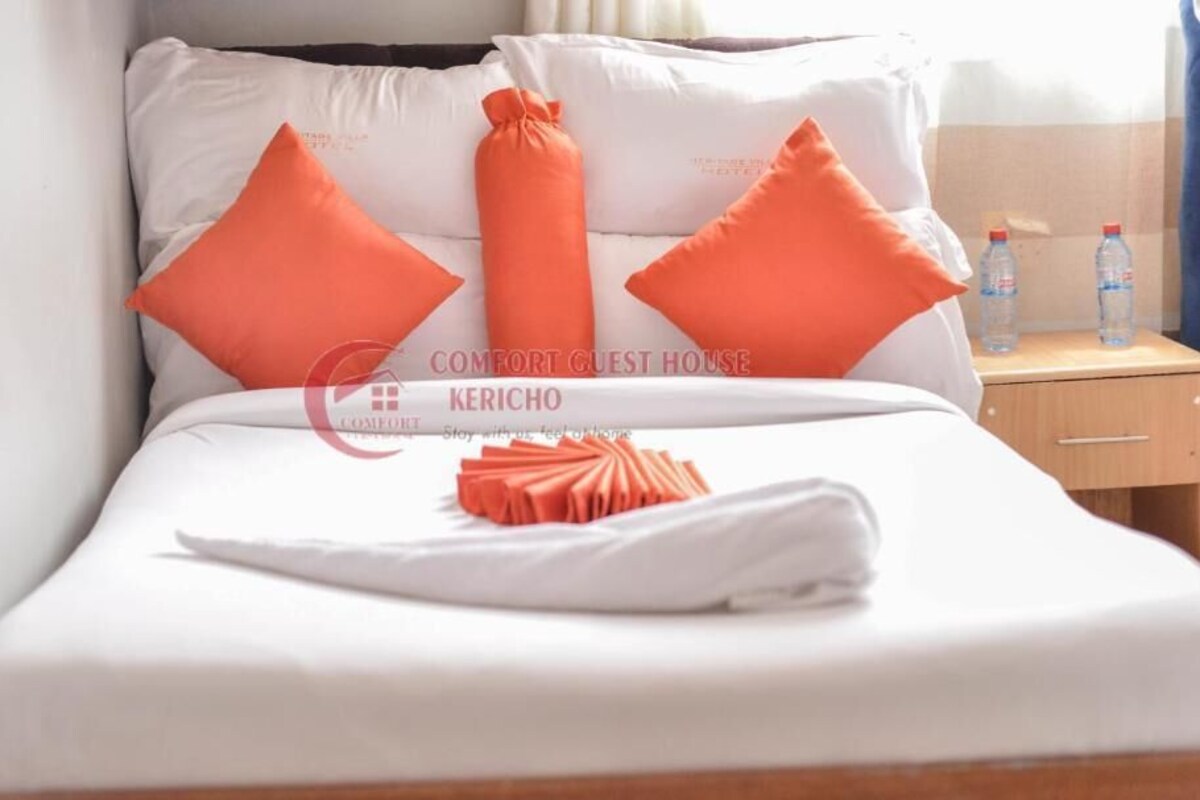 A neatly made bed with white linens and orange pillows, a folded towel, and a wooden nightstand with two water bottles.