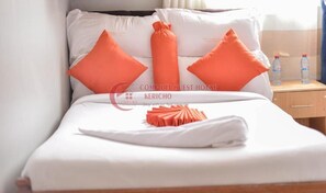 A neatly made bed with white linens and orange pillows, a folded towel, and a wooden nightstand with two water bottles.