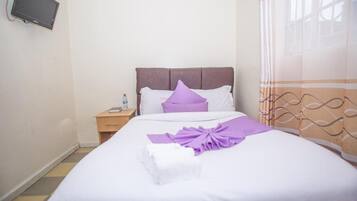 Un lit bien fait, recouvert d’une couvre-lit blanche et violette, avec une tête de lit en bois et une petite table de chevet sur laquelle se trouvent une lampe et une bouteille.