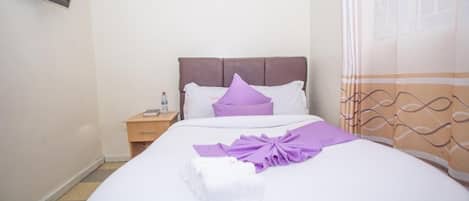 A neatly made bed with a white and purple bedspread, a wooden headboard, and a small bedside table with a lamp and a bottle.