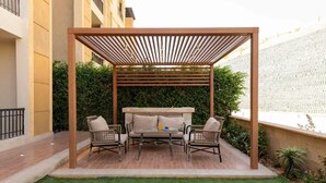 A covered outdoor seating area with a sofa and two chairs, surrounded by greenery and a white fence. - Kennah 3BR with Balcony | Stone Residence, New Cairo - Families Only (New Cairo)