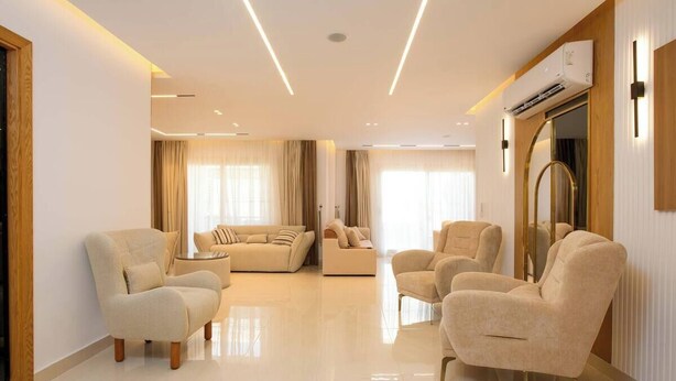 A spacious living room with a large sofa set, two armchairs, and a coffee table. There is a wooden-framed mirror and an air conditioning unit mounted on the wall. - Kennah 3BR with Balcony | Stone Residence, New Cairo - Families Only (New Cairo)
