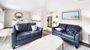 A spacious living room with two dark blue sofas, a round coffee table, and a ceiling fan. A dining area with a white table and chairs is visible in the background. - Heron Marsh Villa 78 (Pawleys Island)