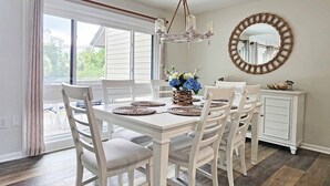 A dining room with a white table and chairs, a chandelier, a mirror, and a view of the outdoors. - Heron Marsh Villa 78 (Pawleys Island)