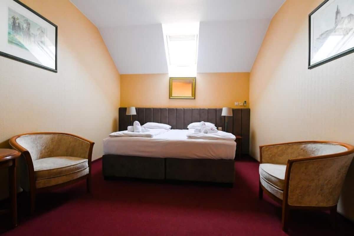 A hotel room with a bed, two armchairs, a nightstand, a desk, a window with a view, and a framed picture on the wall.