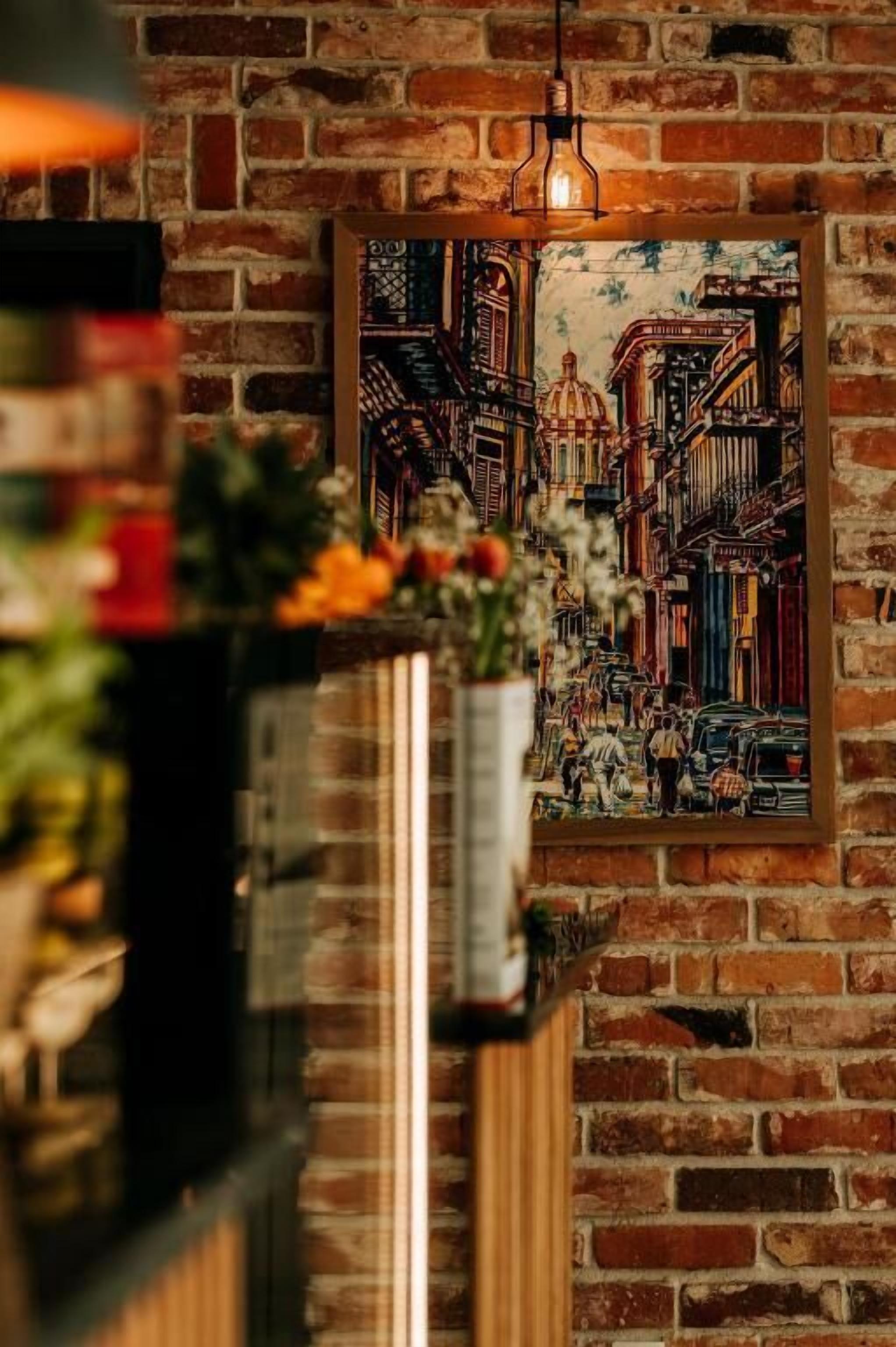 A brick wall with a framed painting of a street scene, a hanging light fixture, and a potted plant.