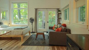 A modern bedroom with a wooden bed, a grey sofa, and a large window offering a view of trees. - Nature view cabin in the pines w/ full bath - 05 (Freeport)