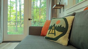 A modern living room with a gray sofa, a window with a view of trees, and a wall-mounted shelf. - Nature view cabin in the pines w/ full bath - 05 (Freeport)