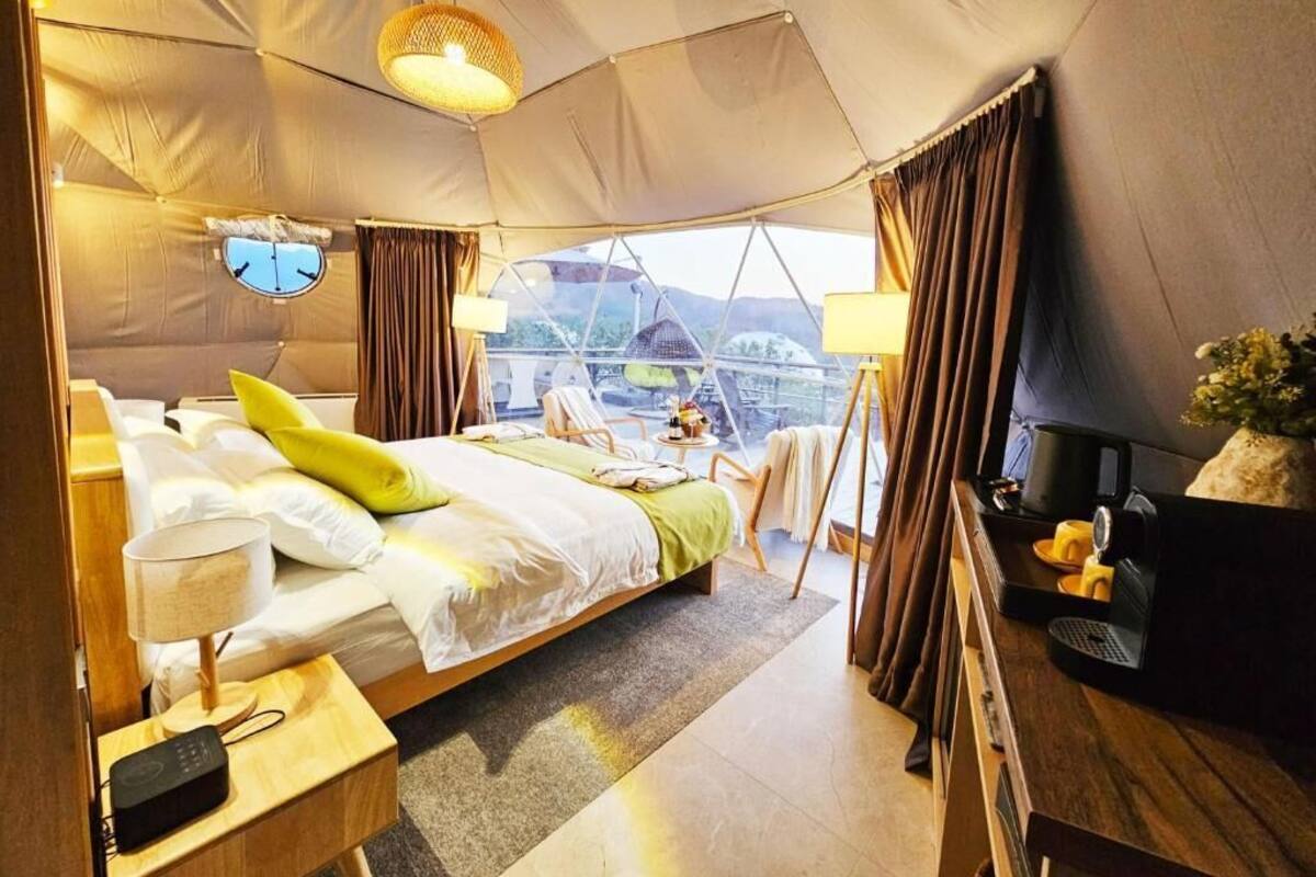 Habitación tipo tienda con cama, mesita de noche, lámpara y vistas al exterior.