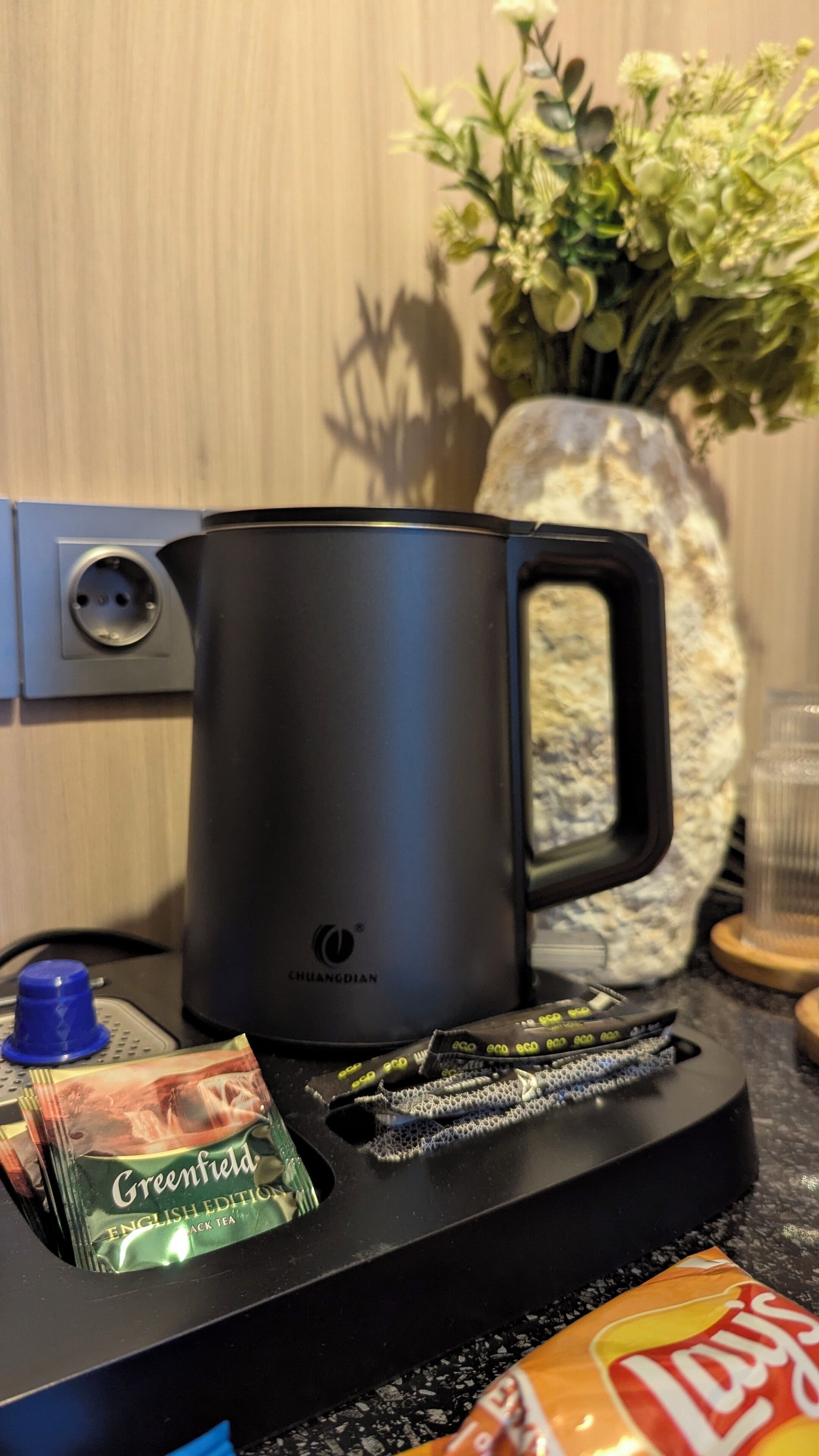 A kitchen area with a black electric kettle, a packet of Green Tea, and a packet of Lays chips on a countertop.