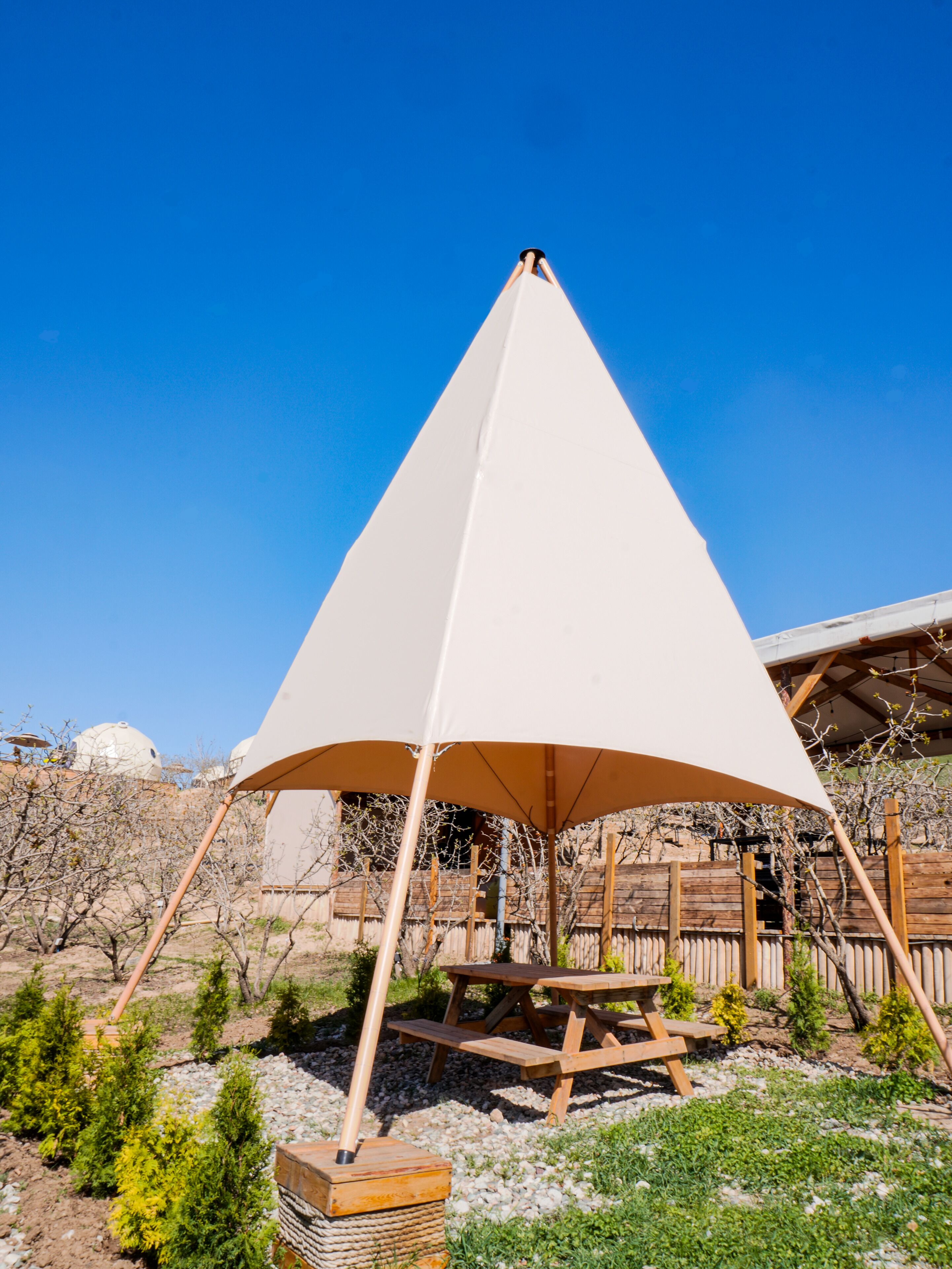 A conical tent structure with a picnic table underneath, set in a landscaped garden with small trees and a wooden fence in the background.