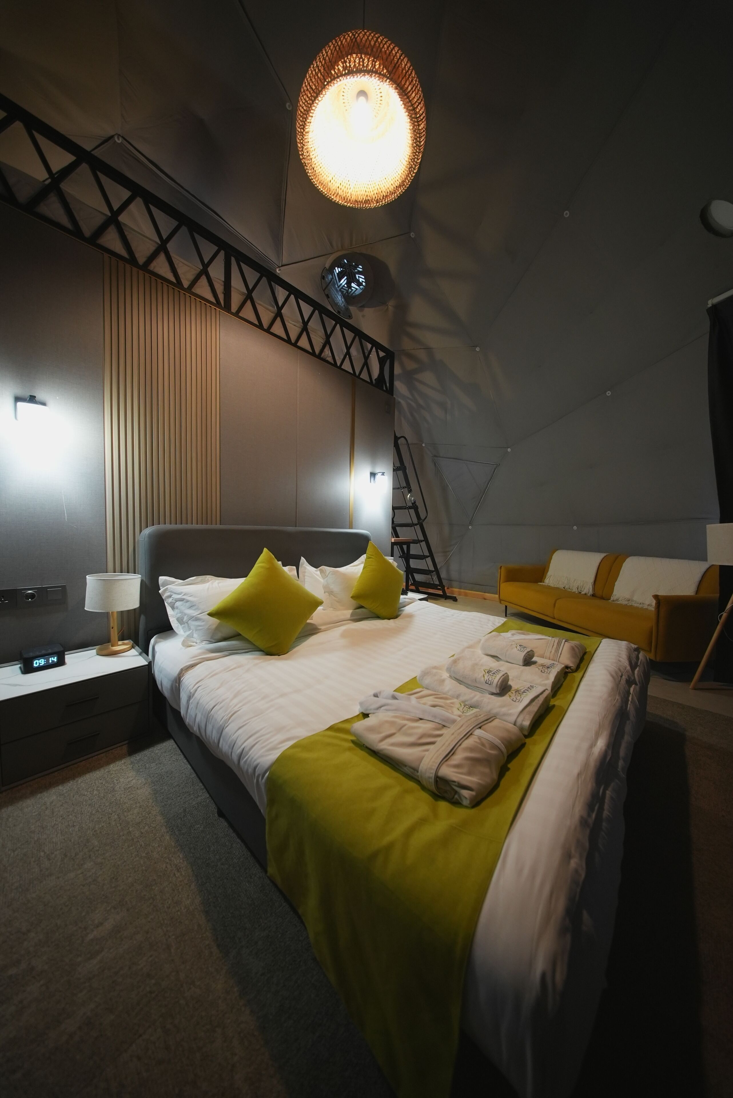 A modern hotel room with a bed, bedside tables, a lamp, a sofa, and a unique ceiling light fixture.