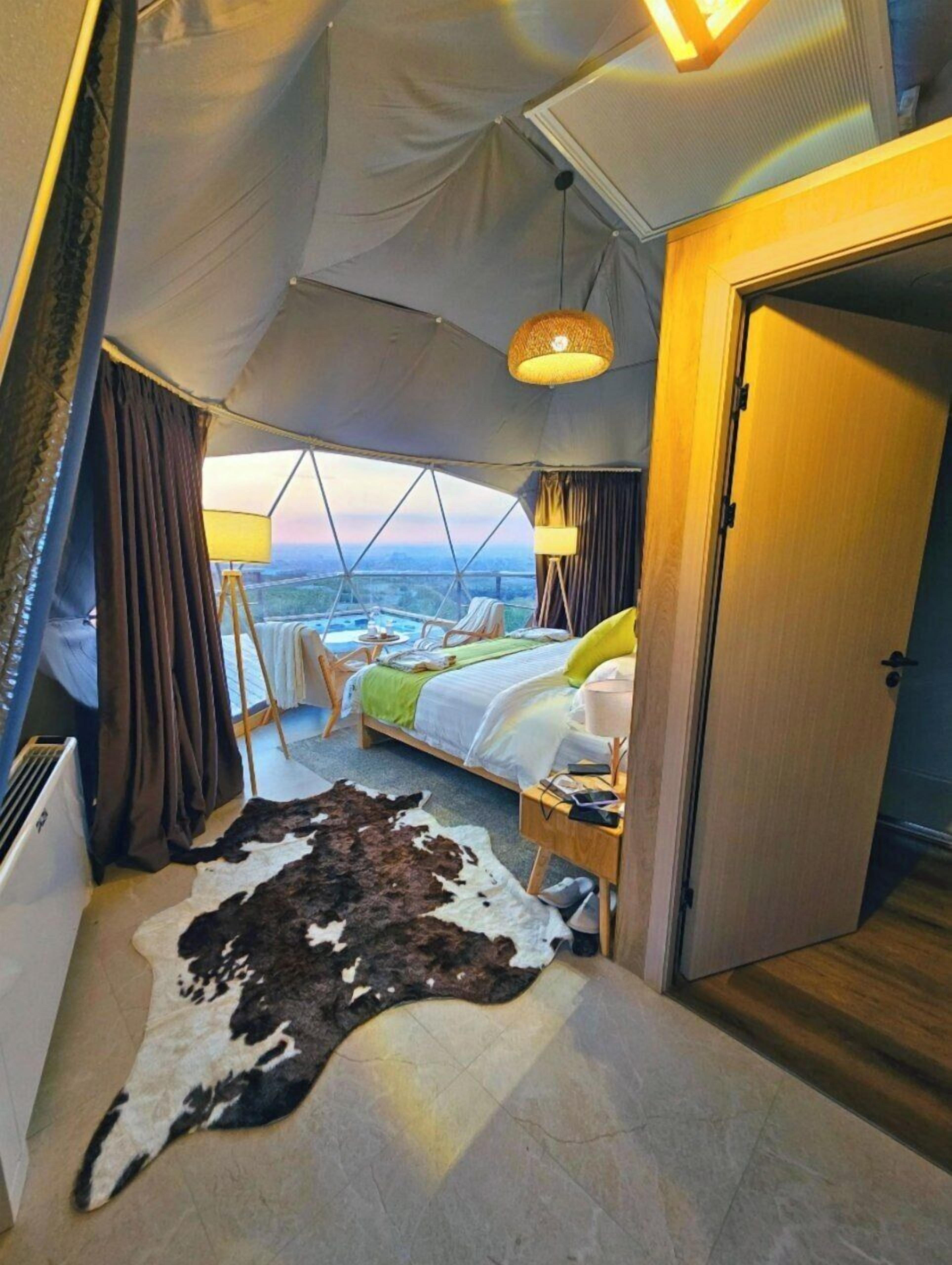 A tented room with a bed, a table with chairs, a lamp, and a view of the landscape.