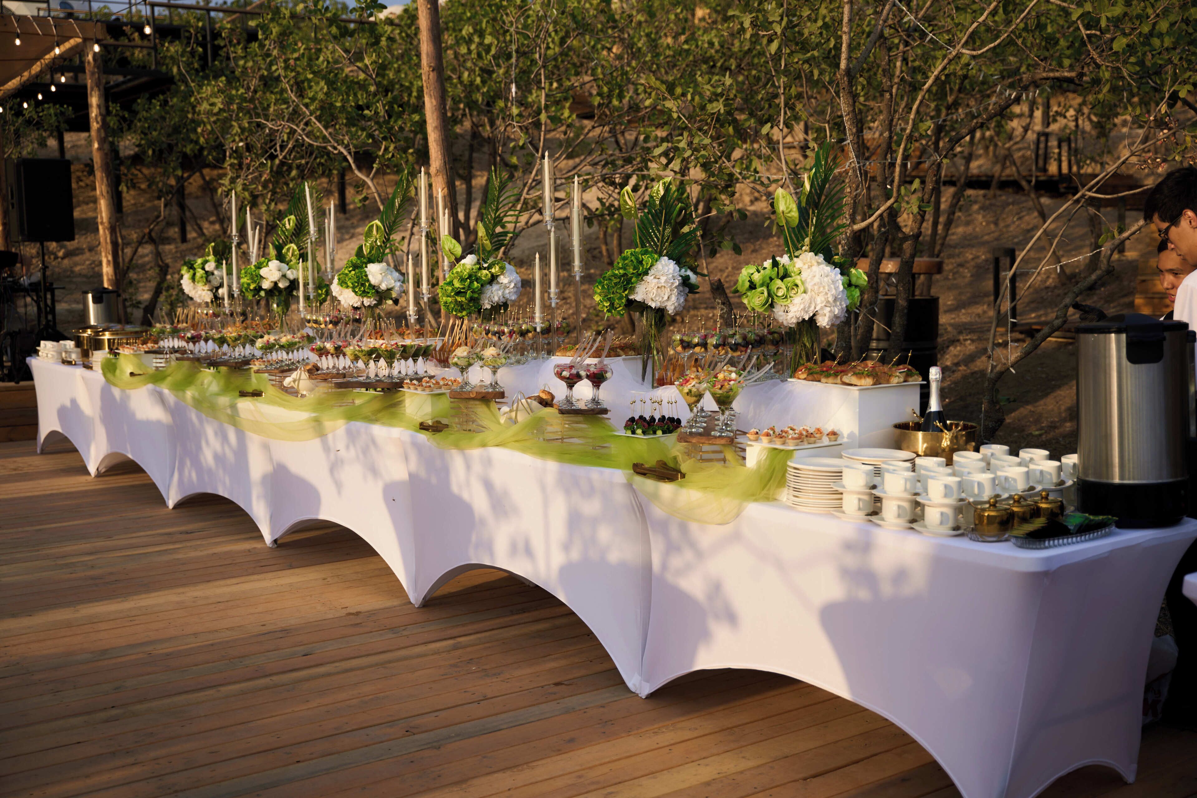 Una larga mesa con mantel blanco, decorada con flores y diversos alimentos.