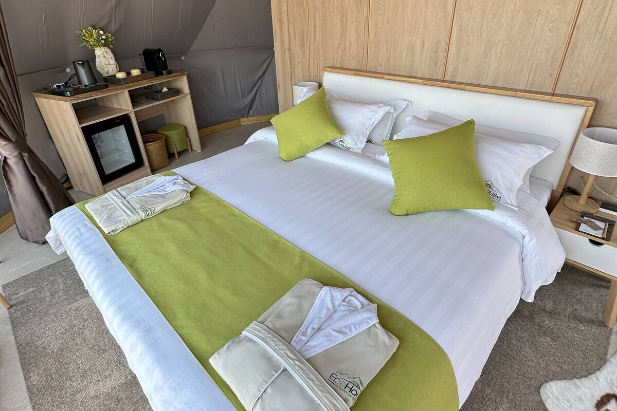 Una cama bien hecha con una colcha verde y blanca, dos almohadas y un cabecero. Una mesita de noche de madera con una lámpara y una pequeña mesa con un florero y velas está al lado de la cama.
