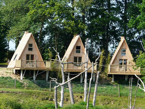 Trois cabanes en bois dotées de balcons, entourées d’arbres et bordées d’une zone herbeuse.