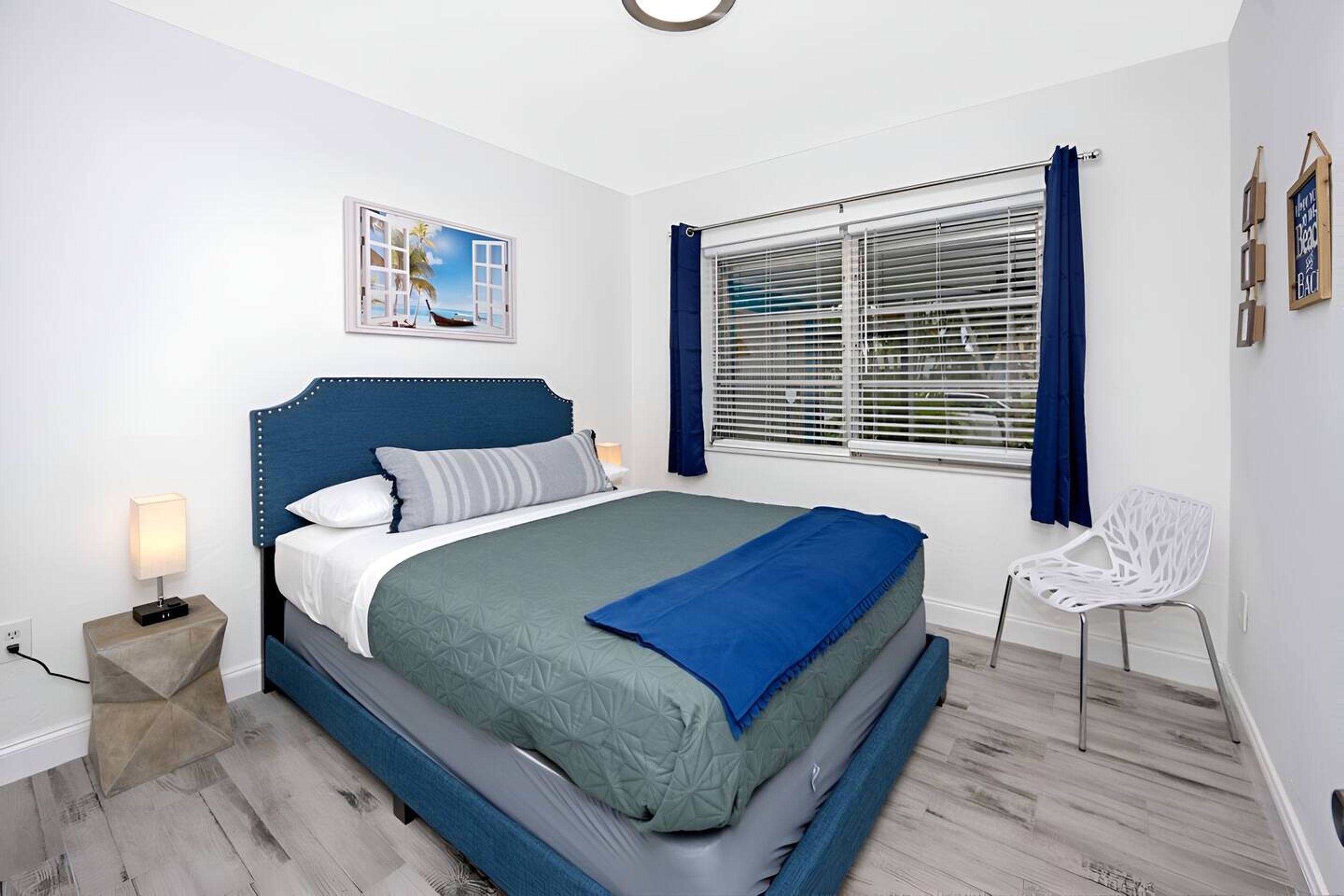 A bedroom with a bed, a bedside table with a lamp, a window with blinds, and a chair.