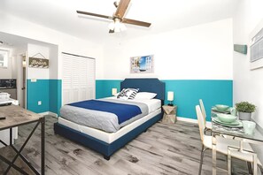 A bedroom with a bed, a desk, a chair, a ceiling fan, and a small table with a plant.