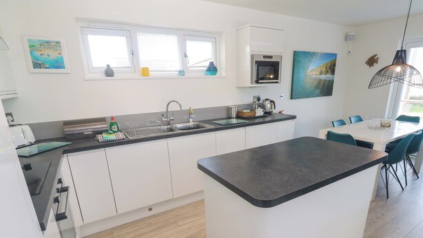 A modern kitchen with white cabinets, a black countertop, and a built-in oven. There is a dining area with a white table and teal chairs. - Lodge 83, Gwel an Mor (Redruth)
