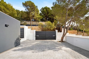 A paved driveway leading to a gated entrance, surrounded by white walls and trees.
