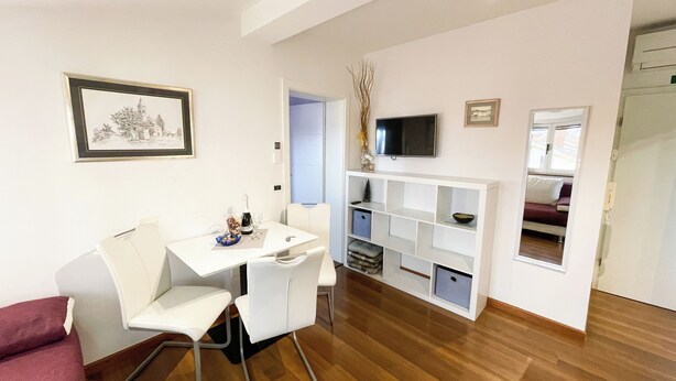 A modern living room with a white sofa, a dining table with chairs, a TV mounted on the wall, and a wooden floor.