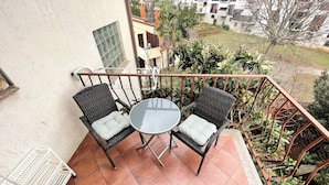 A balcony with two wicker chairs and a small table, surrounded by a metal railing and potted plants.
