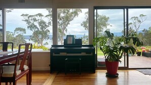 Una habitación con un piano, una mesa de comedor con sillas y un amplio ventanal con vistas a árboles y montañas.