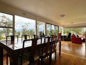 A dining area with a long table and chairs, large windows with a view, and a red sofa in the background.