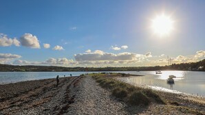 A pebbled beach with a clear sky and a few clouds, a calm body of water, and a few boats.