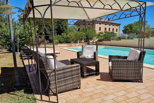 A covered outdoor seating area with wicker furniture, a pool, and a building in the background.