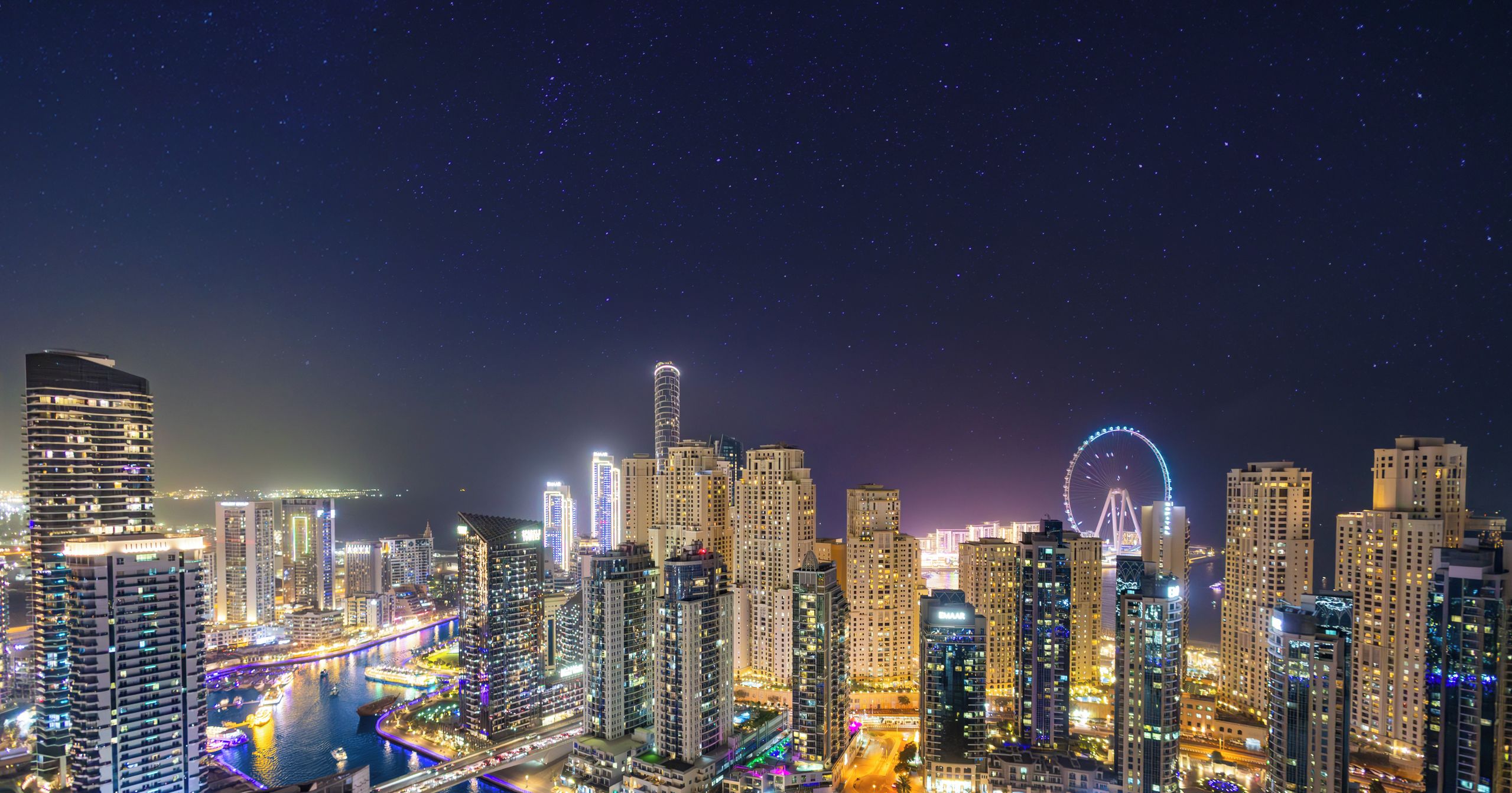 Une vue nocturne d’une ville, avec des bâtiments illuminés et une grande roue, ce qui suggère un cadre urbain luxueux.