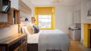 A bedroom with a bed, a window with yellow blinds, a post office sign, and a kitchen area.