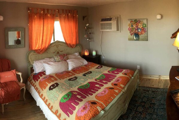 A bedroom with a bed, a chair, a nightstand, a wall-mounted air conditioner, and a painting.