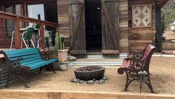 A rustic wooden building with a covered porch, two benches, a fire pit, and a cactus plant.