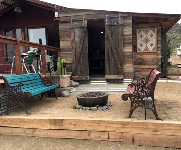 A rustic wooden building with a covered porch, two benches, a fire pit, and a cactus plant. - Romantic Yucca Valley Cabin with Hot Tub (Yucca Valley)