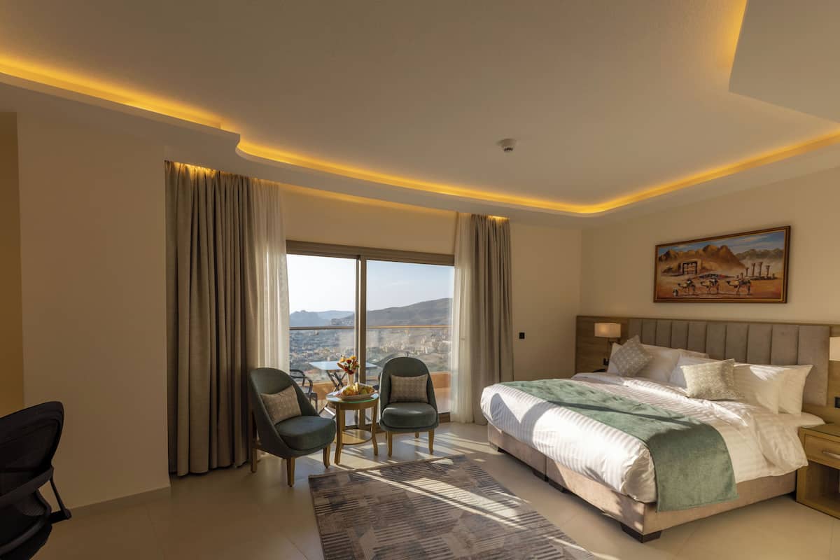 A hotel room with a large bed, two armchairs, a small table with a vase, and a painting on the wall.