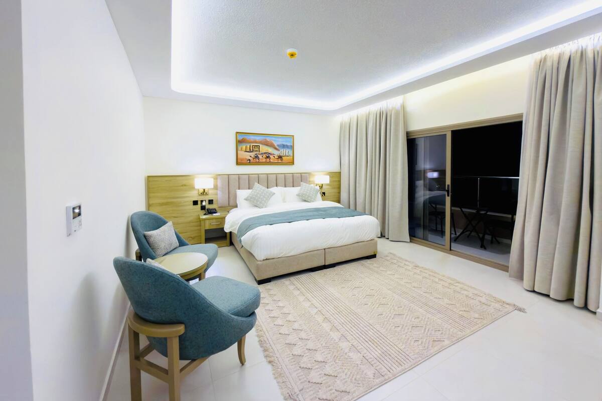 A hotel room with a bed, a desk, a chair, a painting, and a sliding glass door to a balcony.