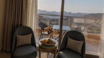 Een kamer met twee stoelen, een kleine tafel met een schaal fruit en een uitzicht op een stadsbeeld door grote ramen.