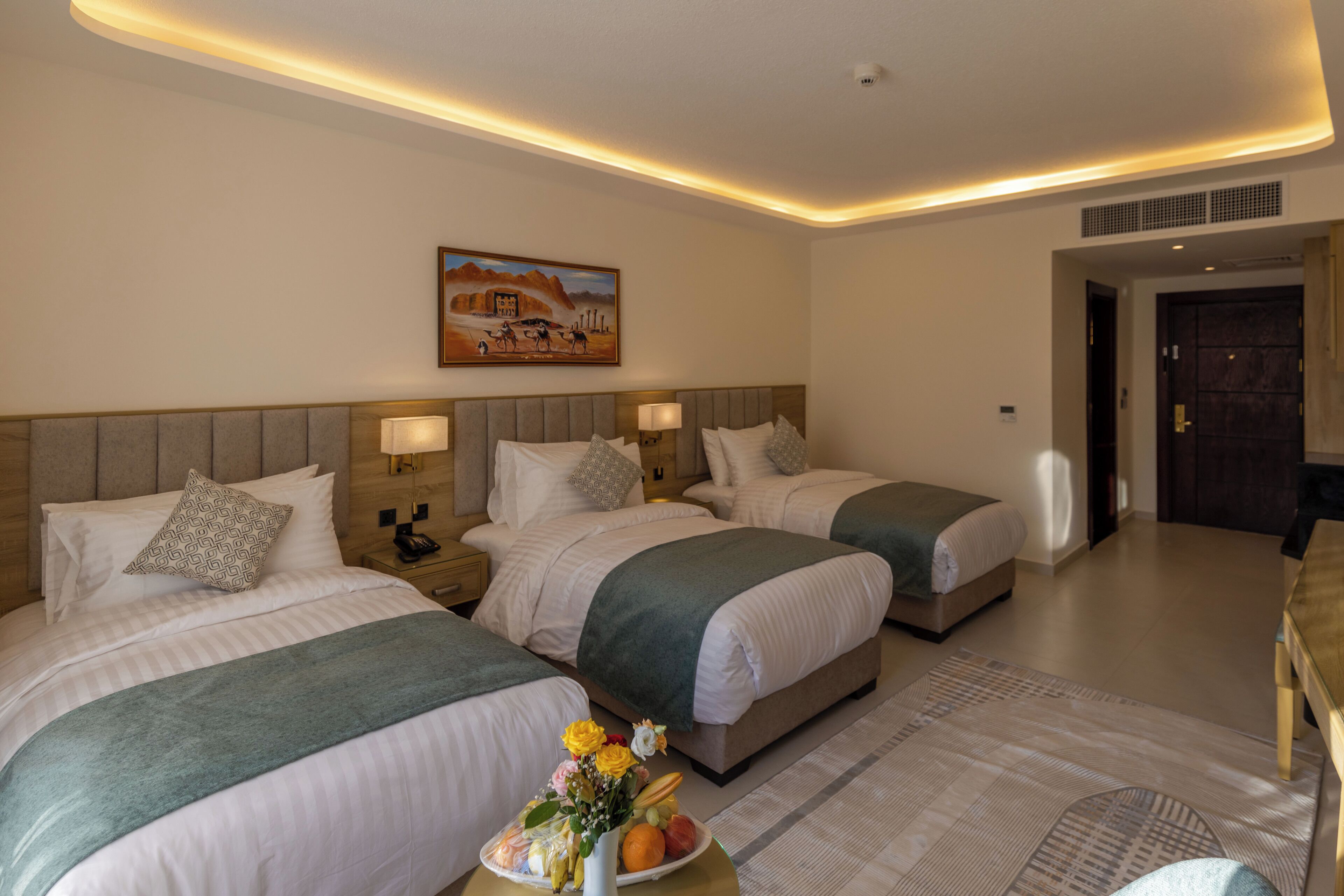 A hotel room with three beds, a painting on the wall, and a small table with fruit and flowers.