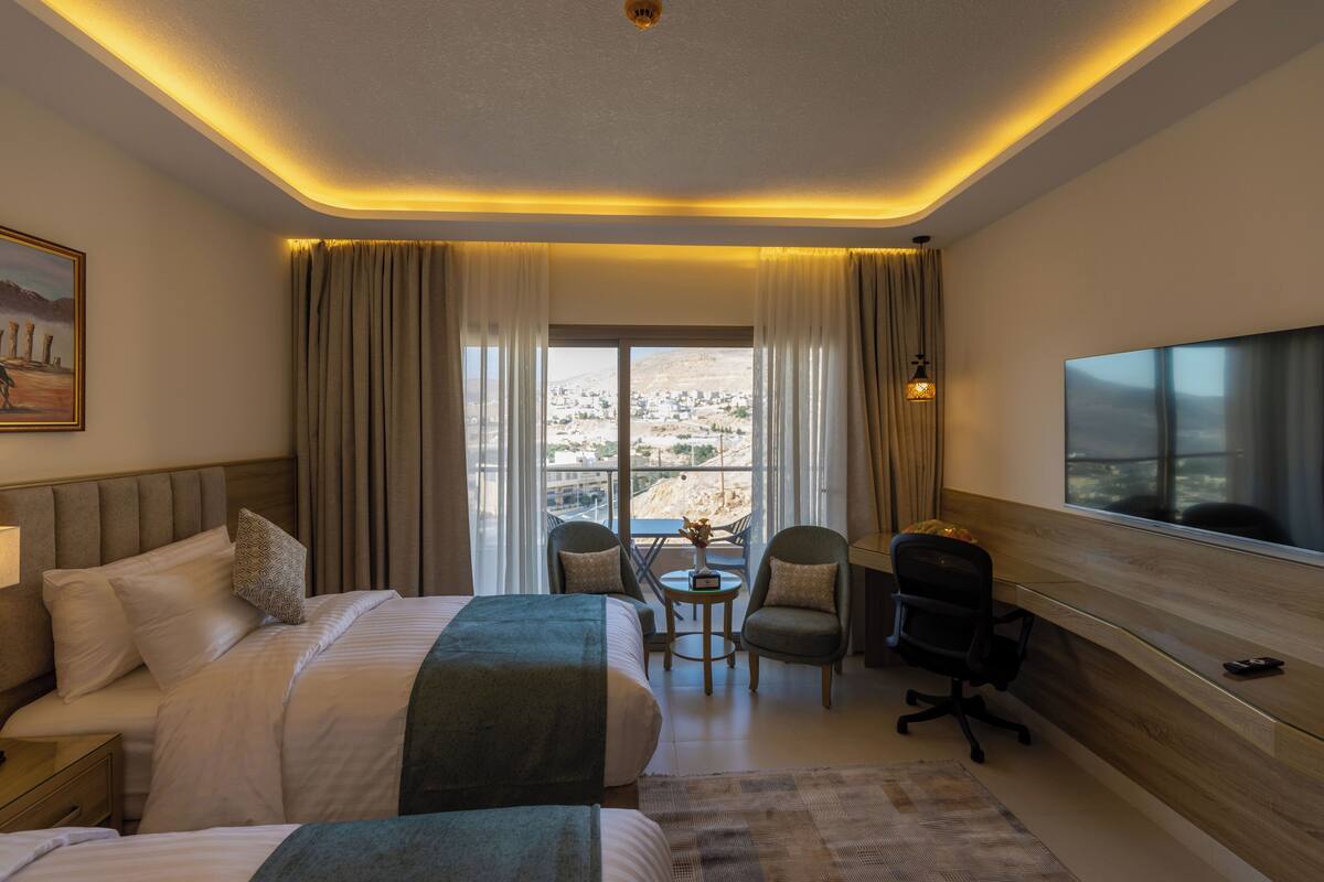 A hotel room with a large bed, a desk, two chairs, a TV, and a view of the city through the window.