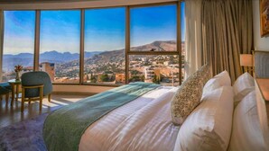 Ein Schlafzimmer mit großem Fenster und Blick auf die Stadt, einem Bett mit gemusterten Kissen und einem kleinen Tisch mit Stuhl.