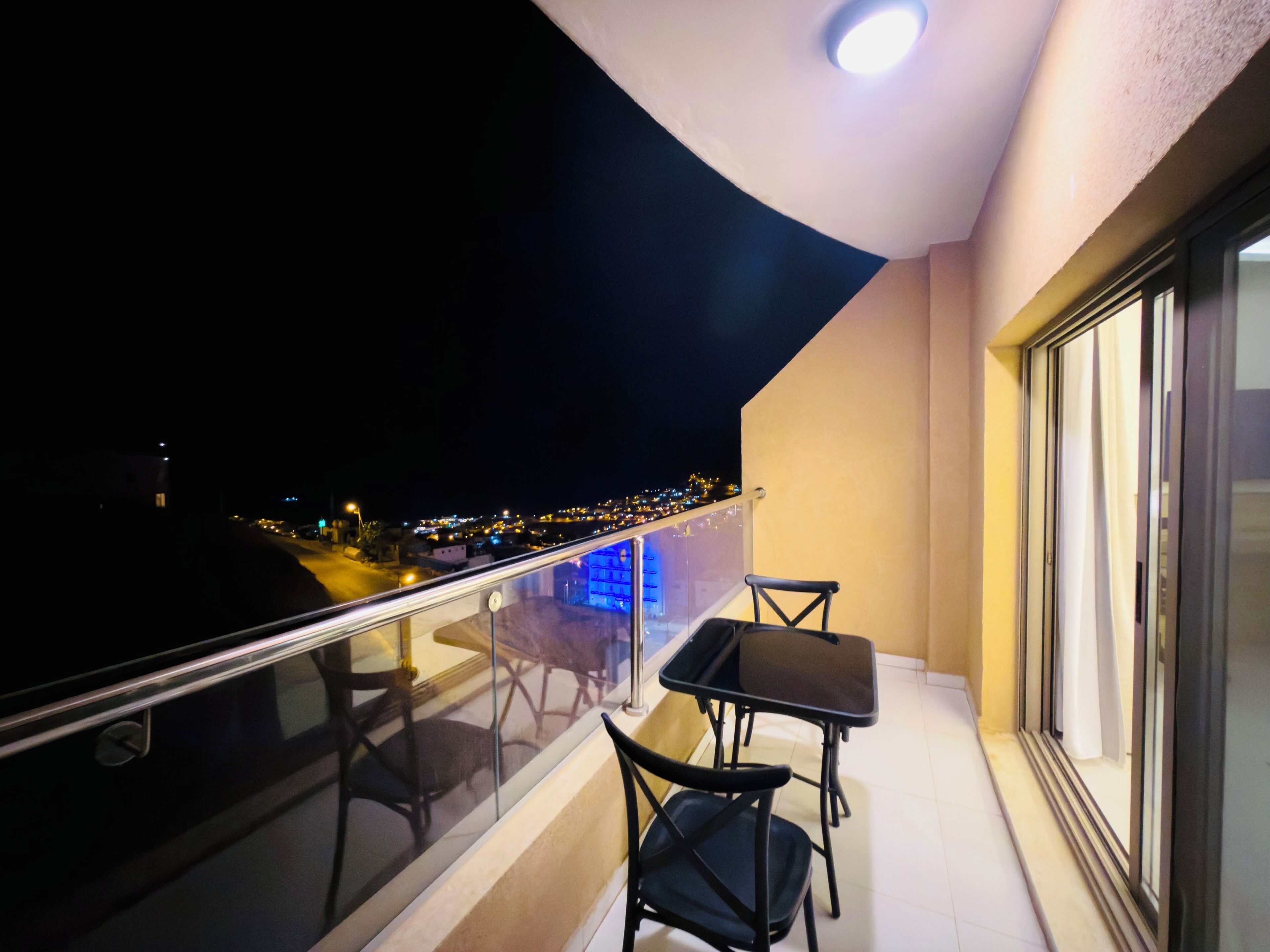A balcony with a glass railing, two chairs, a small table, and a view of a cityscape at night.