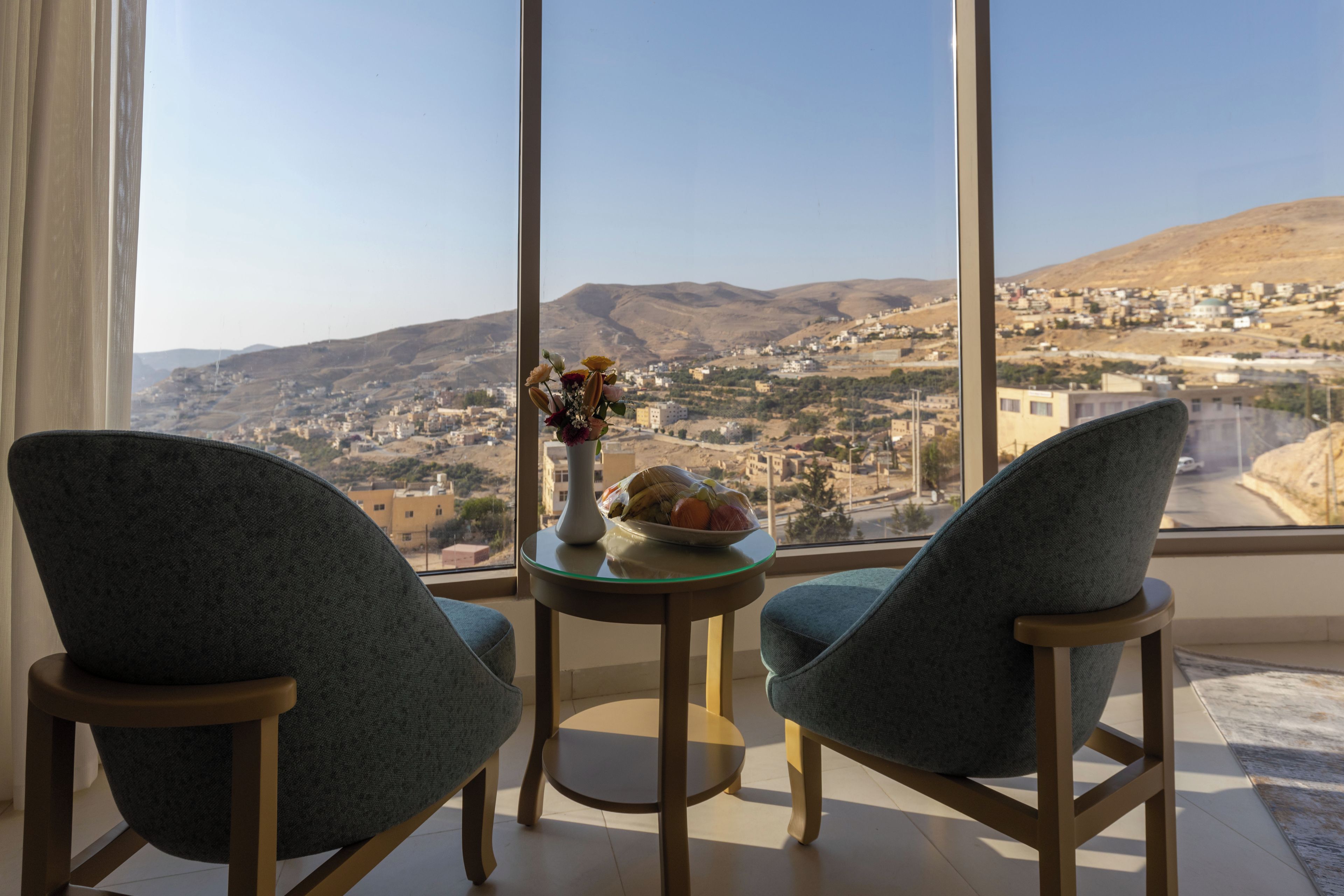 A room with two chairs facing a table with a vase of flowers and a plate of fruit. A large window offers a view of a town and hills.