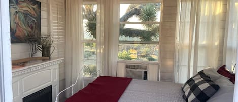 A bedroom with a bed, a fireplace, a window with curtains, and a view of the outdoors.