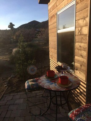A wooden building with a patio table and chairs, a window, and a view of a hilly landscape. - Unique Yucca Valley Rental Ideal for Romantic Getaways in California (Yucca Valley)