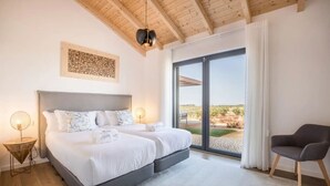 Ein Schlafzimmer mit einem großen Bett, zwei Nachttischen mit Lampen, einem Sessel und Blick ins Freie.