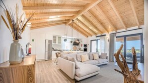 Ein modernes Interieur mit Holzbalkendecke, einem Wohnbereich mit Sofa, einer Küche und Blick durch große Glastüren.
