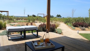 A wooden deck with two lounge chairs, a table with a tray of food and drinks, and a view of a vineyard and ocean.