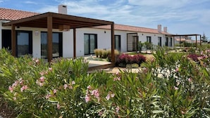 A modern building with a covered patio area, a garden with flowering plants, and a clear blue sky.