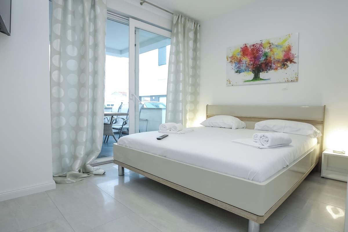 Una cama bien hecha con sábanas blancas, un control remoto, un cuadro de un árbol colorido en la pared y vistas al exterior a través de una puerta de cristal.